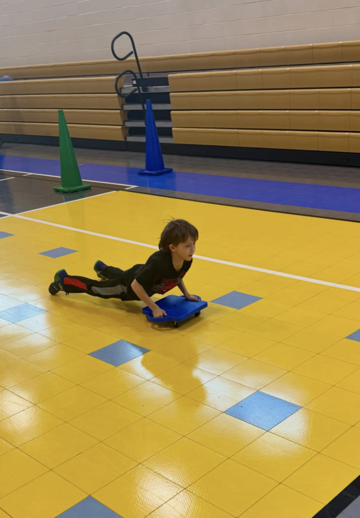 A student holds on to a rolling platform and glides across a school gym floor.