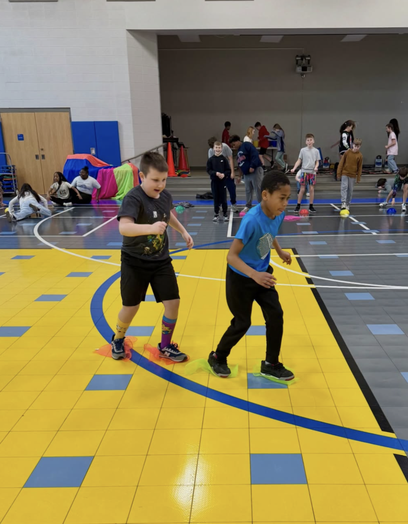 Image is of a school gym with students inside. Two students at the front of the image race across the gym floor while standing on bright colored cloths.