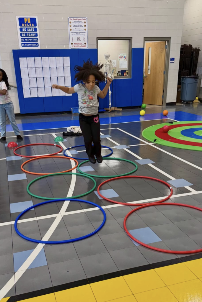 A student jumps into the middle of hula hoop that's sitting on a school gym floor.