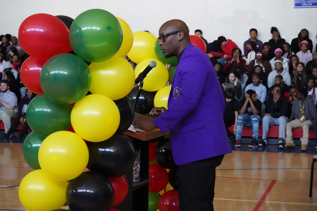 Erwin Middle School's principal speaks into a microphone inside the school gym. He stands at a podium surrounded by red, green, yellow and black balloons. Students sit in the bleachers behind him.