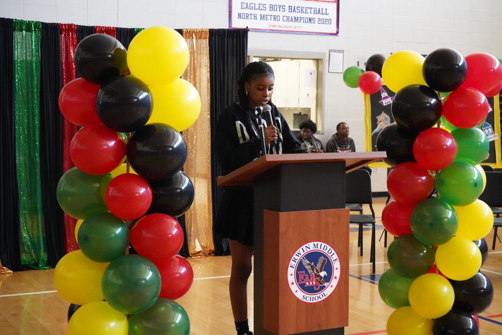 A girl speaks into a microphone during the Black History Program held in the Erwin Middle School gym. She stands at a podium that's surrounded by black, red, green, and yellow balloons.