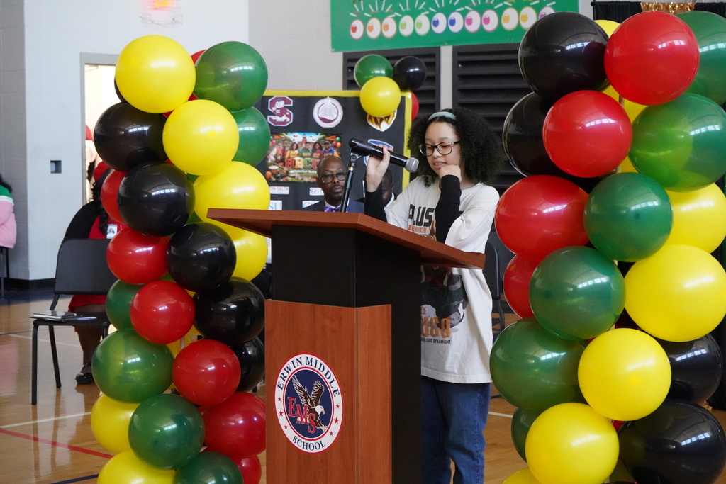 A girl speaks into a microphone during a Black History Program in the Erwin Middle School gym. She stands at a podium that's surrounded by black, red, green, and yellow balloons.