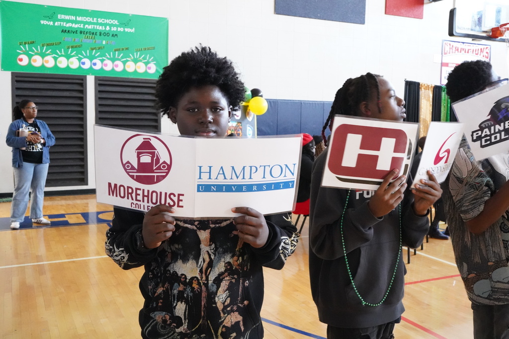 Students stand in a line and hold up signs with logos of HBCUs. The student in the center of the photograph holds signs that say "Morehouse College and Hampton University."
