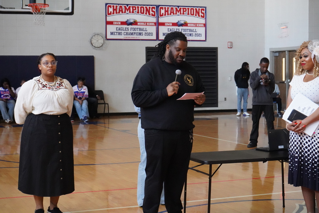 A man speaks into a microphone while performing a short play inside the Erwin Middle School gym. Two women stand next to him.