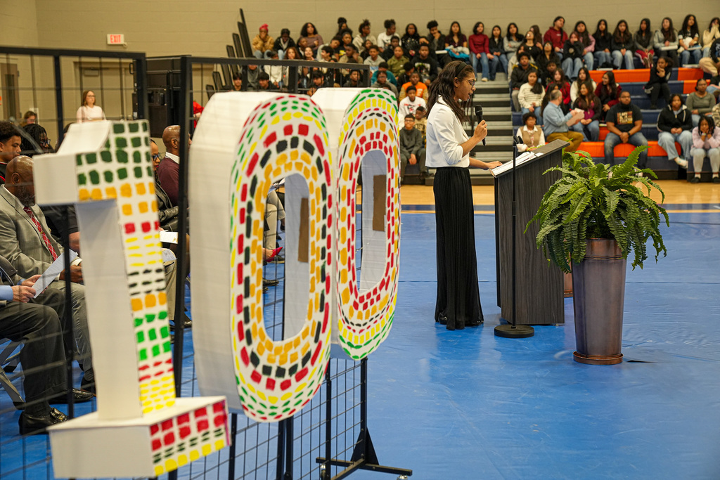 A student delivers a speech during a Black History Month program inside a school gym. Next to her is a large 100 made out of cardboard.