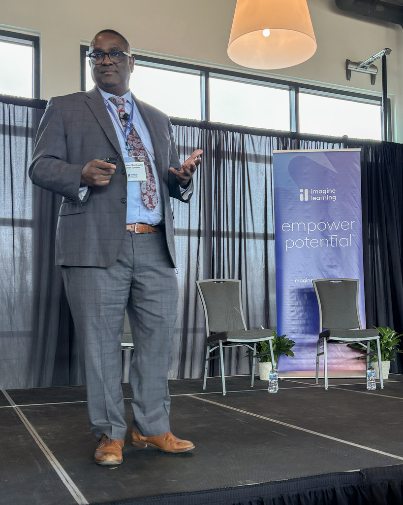 A man delivers a speech on stage. In the background is an Imagine Learning banner that reads. "Empower Potential."