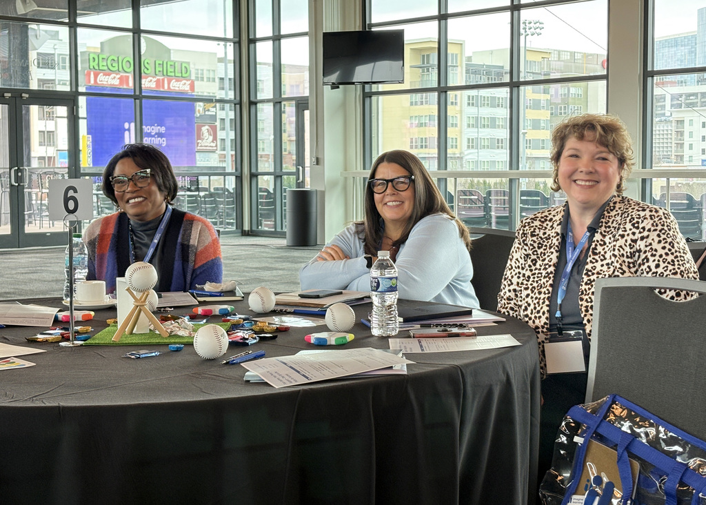 Three women are sitting at a table during a meeting. They are smiling at the camera. 
