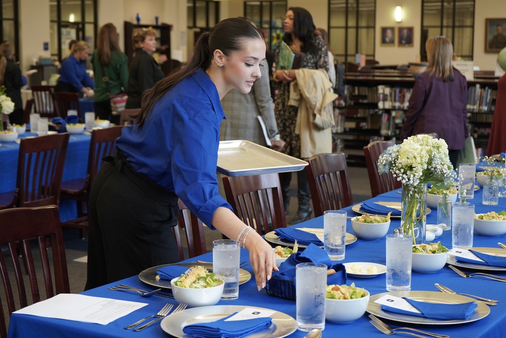 A JEFCOED Culinary Arts student sets bread on a table. The table also has glasses of water, salad, and flowers on it.