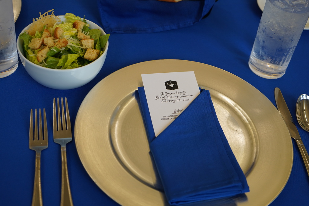 A folded napkin with a menu sits on top of a gold plate. Forks, a spoon, a knife, a salad bowl and glasses of water sit nearby. All of these items are on a table with a blue table cloth on it.