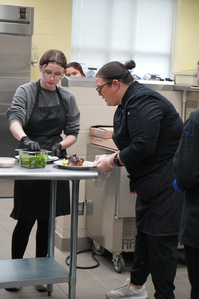 A JEFCOED Culinary Arts student works inside a kitchen. An educator stands next to her and holds a plate.