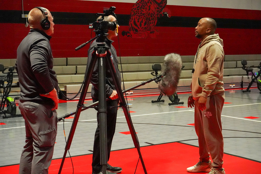 Three men stand inside a school gym. One man operates a camera while another man holds a microphone. The third man speaks into the microphone.