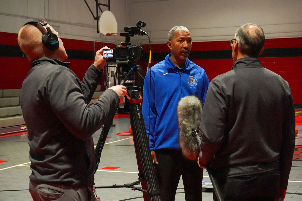 Three men stand inside a school gym. One man operates a camera while another holds a microphone. The third man speaks into the microphone.