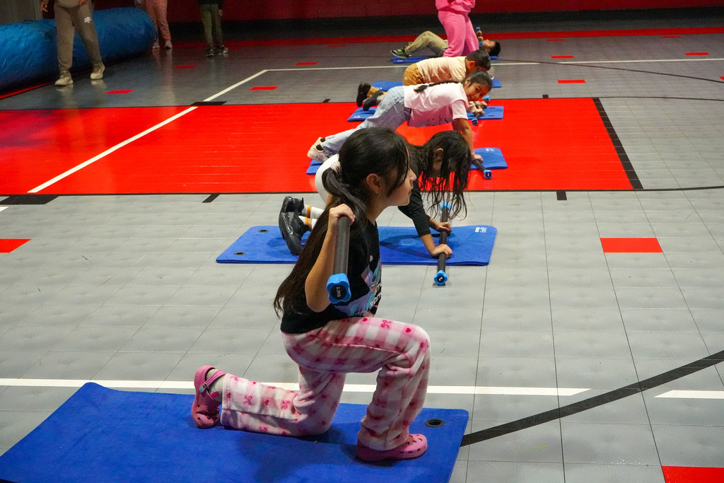 A student does a lunge with a training bar inside a school gym. In the background, other students perform exercises on workout mats.