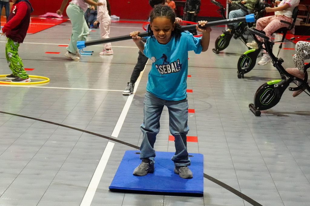 A student performs an exercise while holding a training bar. Other students complete exercises behind him.