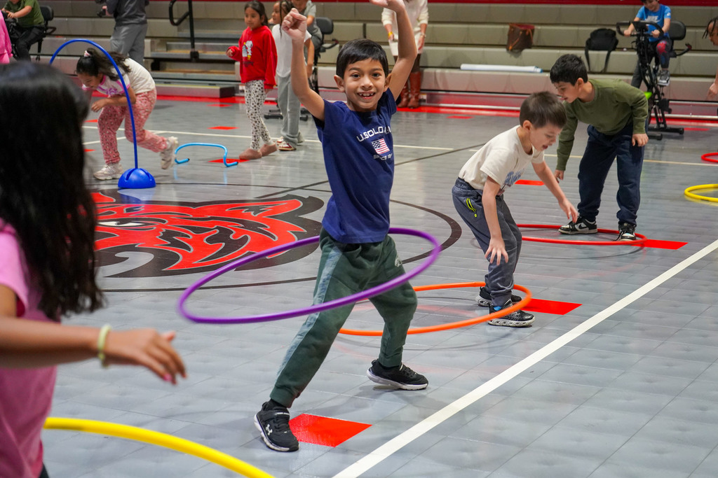Students hula hoop while inside a school gym. In the background, other students run through hurdles.