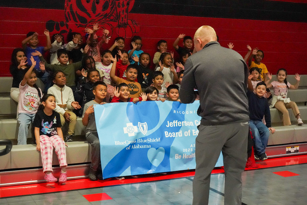 Students sit on bleachers inside a school gym and smile and wave. A man holding a video camera takes video of them. The students are also holding a sign that recognizes Jefferson County Schools as the recipient of a Blue Cross Blue Shield of Alabama Be Healthy Grant.