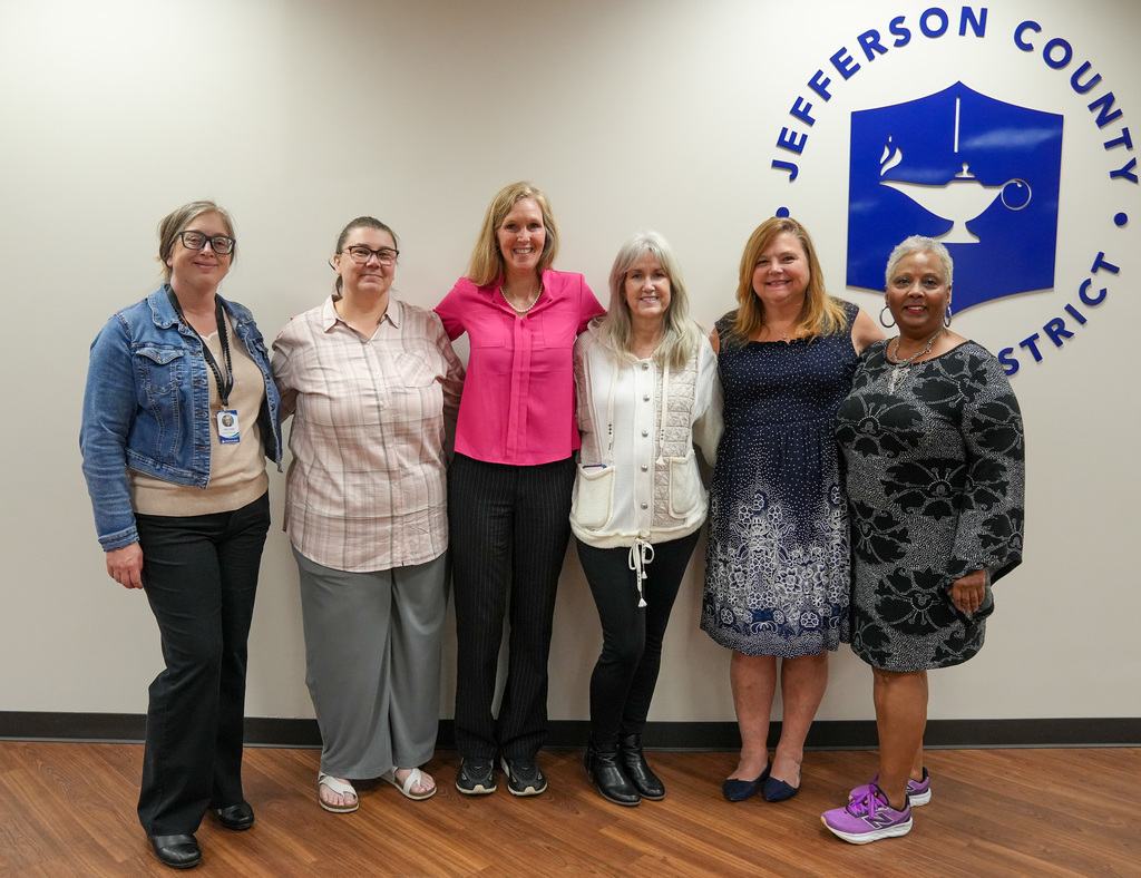 A group of women stand together for a photo inside a room and smile. They stand in front of a wall with a  large sign displaying the Jefferson County Schools logo.