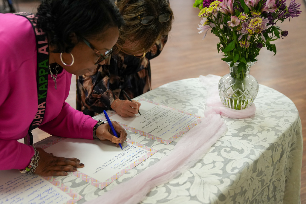 Two women sign cards that sit on a table. Beside the cards is a vase with flowers inside.