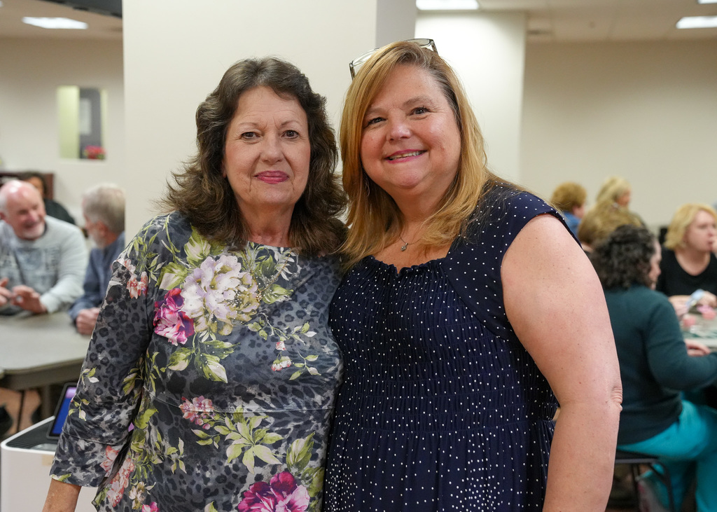 Two women stand together for a photo and smile. One woman wears a shirt with flowers on it and the other wears a navy dress with  dots on it. Behind the women, a group of people sit at tables inside the room.