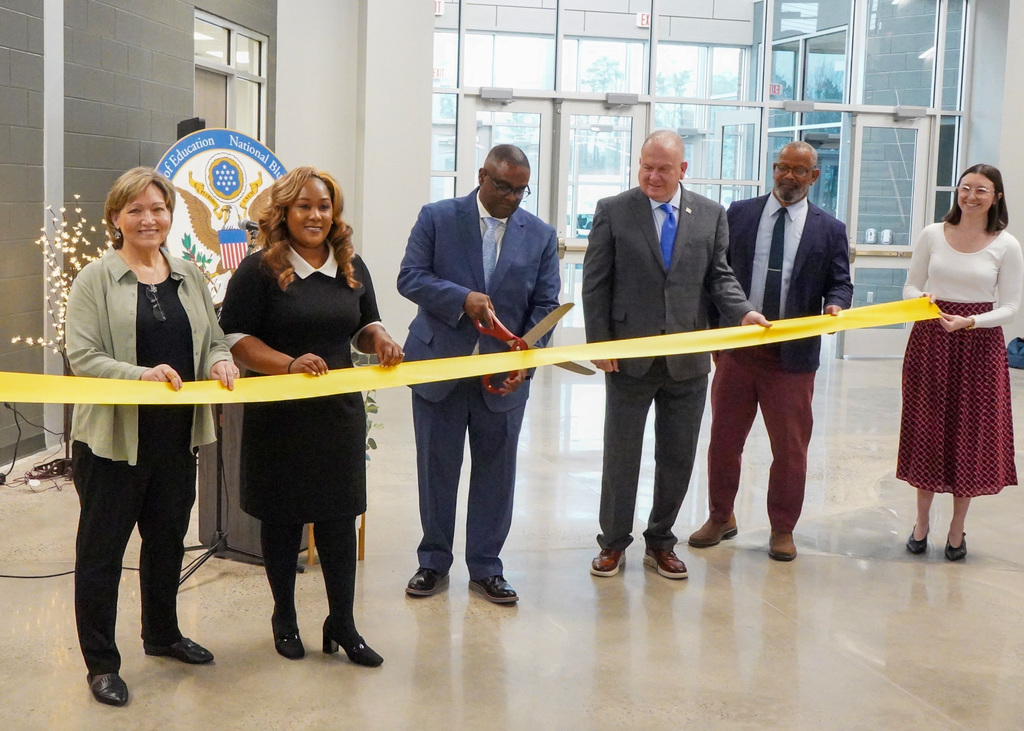 Superintendent Dr. Walter Gonsoulin, board members, and the principal of Jefferson County International Baccalaureate School stand together for a ribbon cutting ceremony. Dr. Gonsoulin cuts the ribbon with large scissors while everyone else holds the ribbon.