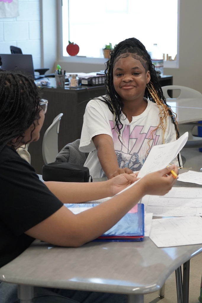 A student smiles while sitting at her desk in a classroom and doing work. A student in front of her also sits at a desk and holds up a piece of paper.