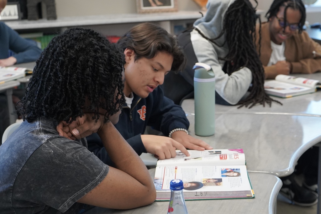 Two students read from a textbook while sitting at desks. Other students behind them do the same.
