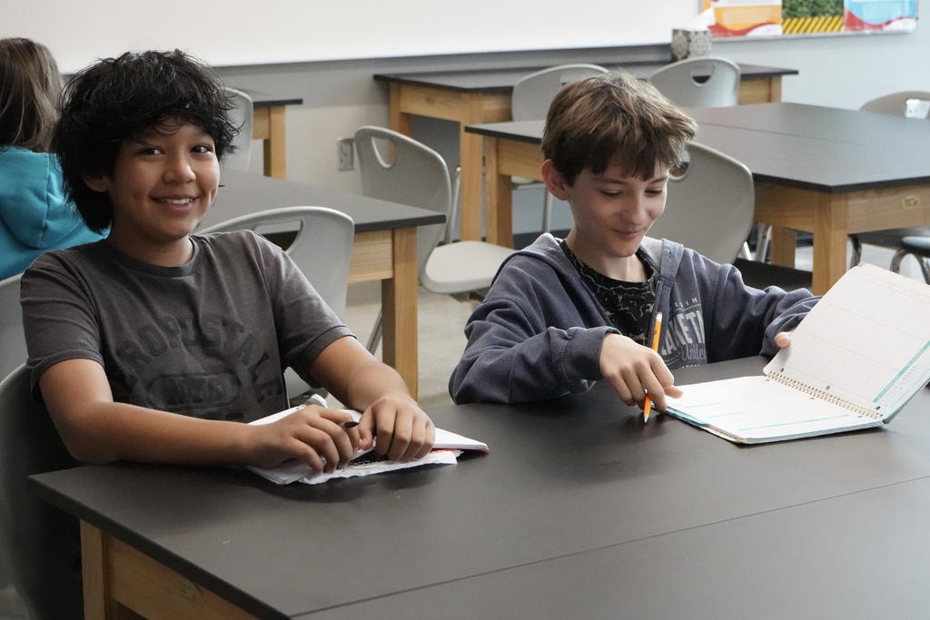 Two students smile while sitting at a table and writing in notebooks.