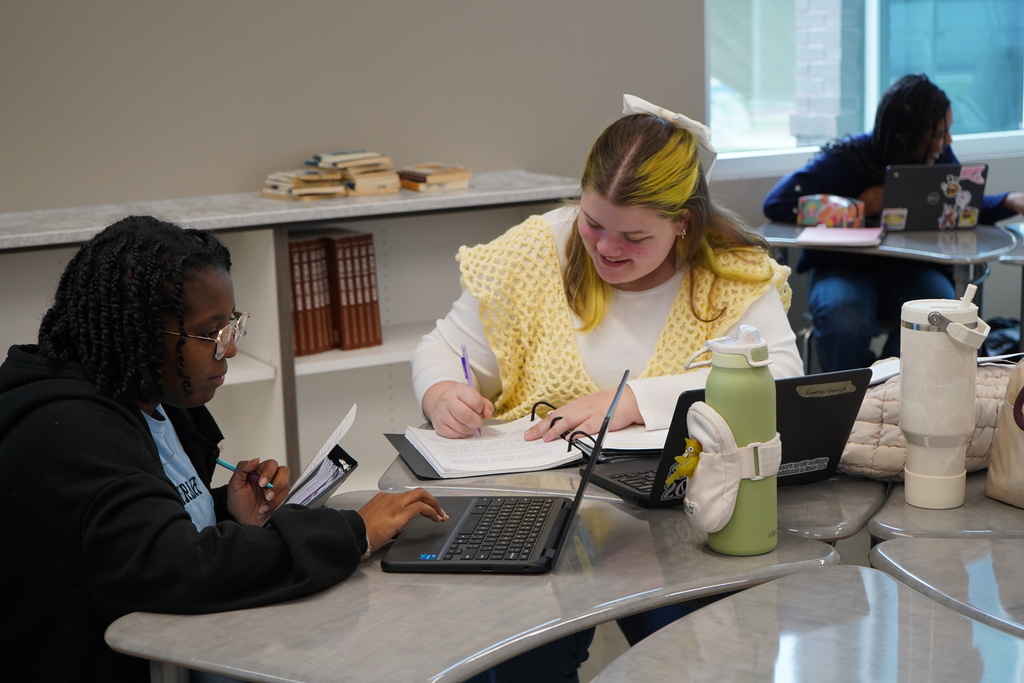 Two students sit at a desks and complete work on paper and laptops.