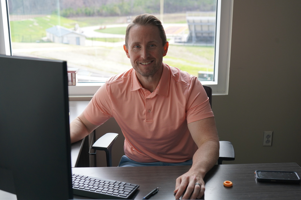 A man sits at a desk while working on a computer and smiles.