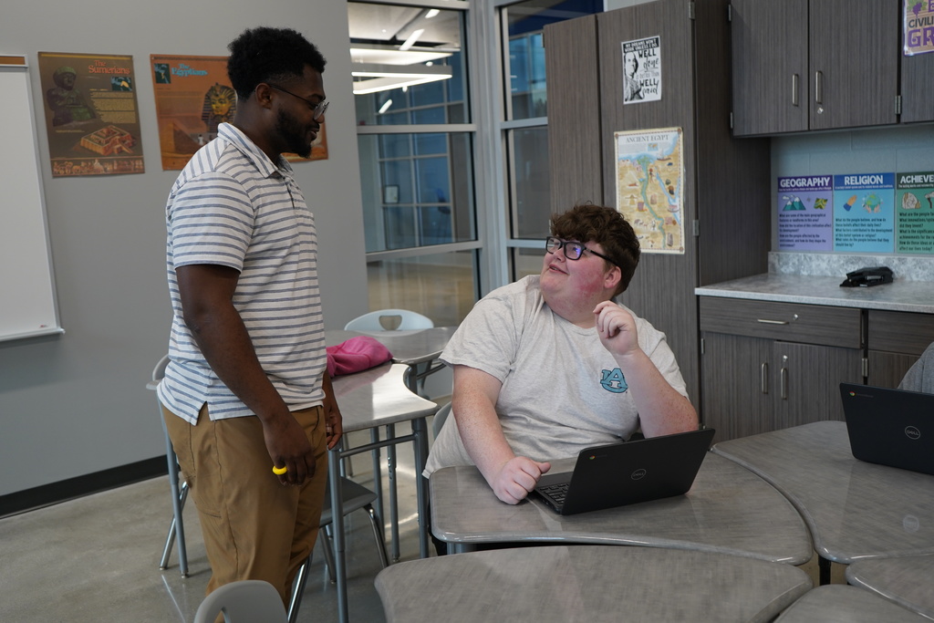 A student smiles while talking to his teacher inside a classroom.