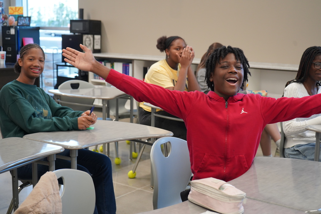 A student sits at a desk in a classroom with other students. The student holds his arms out and smiles. The student behind him also smiles.