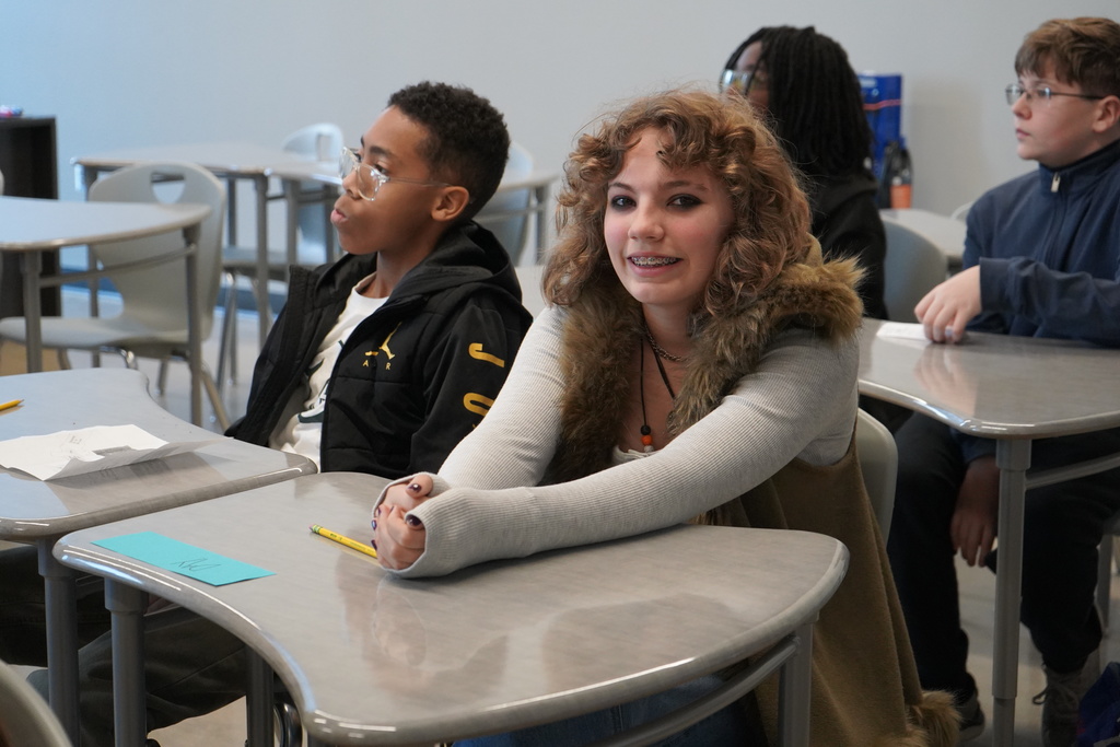 Students sit at desks inside a classroom. One of the students smiles for a photo.