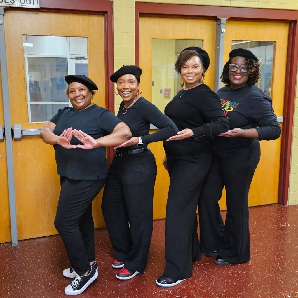 Four African American women wearing matching black shirts and pants as well as a black hat. They are arranged in a horizontal line, each woman sporting a wide smile. The women are making a hand gesture of a triangle shape with their thumbs and pointer fingers connecting.