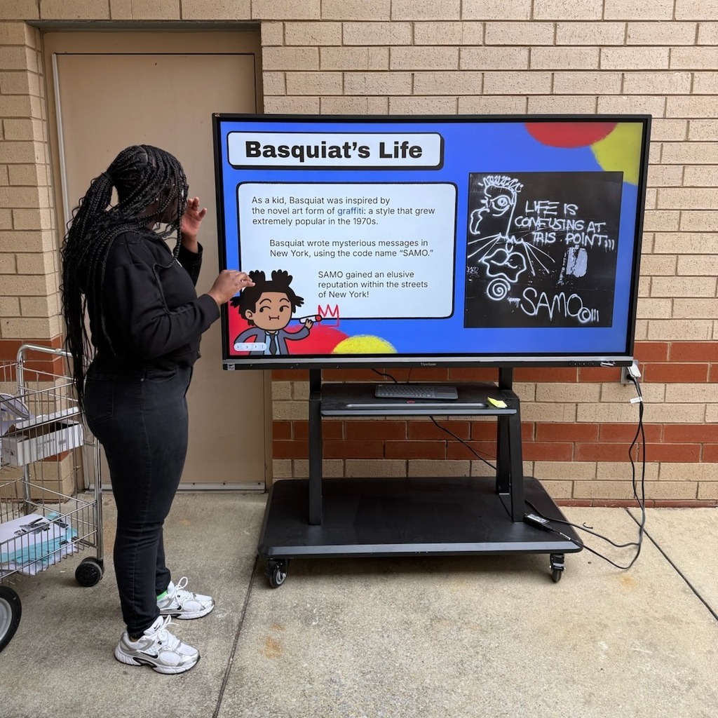 A student is standing in front of a presentation on a tv screen that reads, "Basquiat's Life."