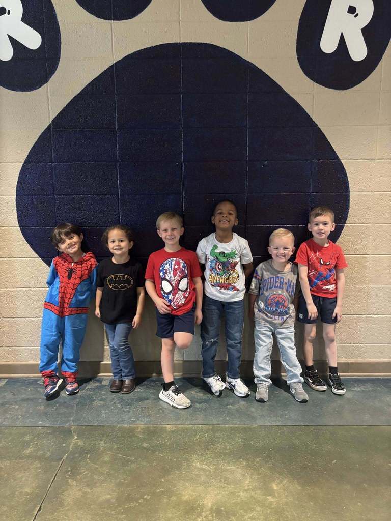 A group of young students stand together for a photo. They wear super hero themed shirts and outfits.