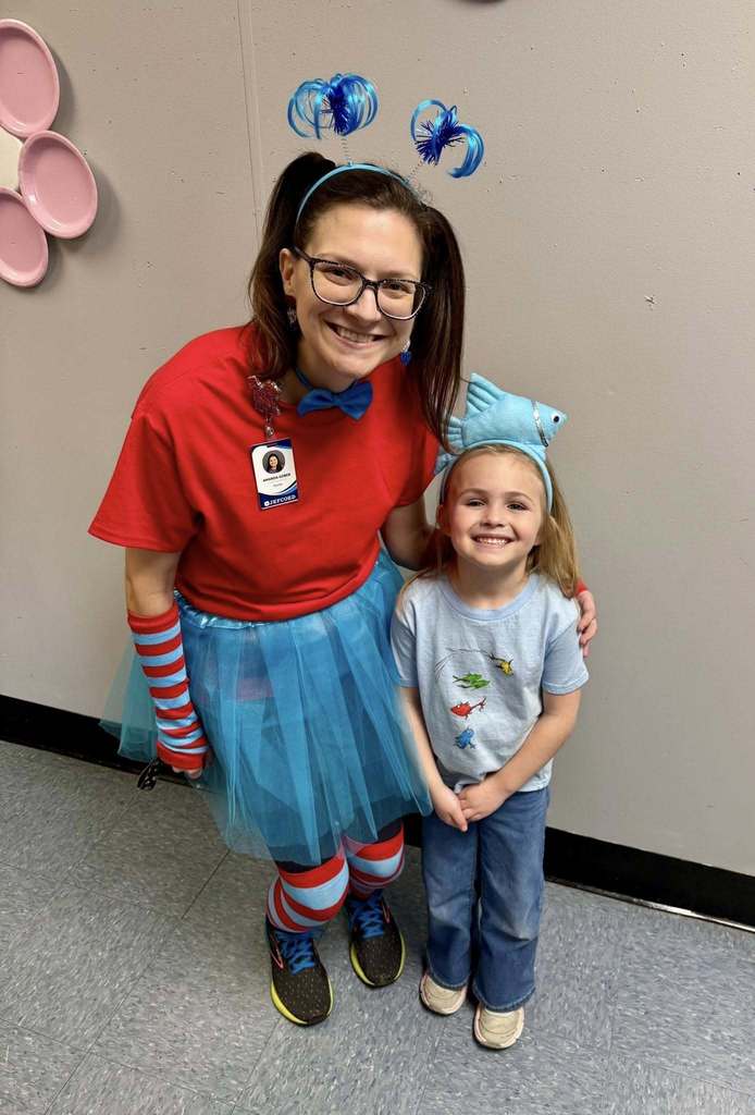 An adult wearing a Dr. Suess themed outfit poses for a photo with a little girl. The girl has a headband with a fish on it and a shirt with fish on it.