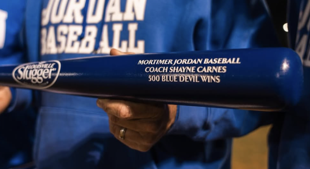 A man holds a bat that says "Mortimer Jordan Baseball Coach Shayne Carnes 500 Blue Devil Wins"