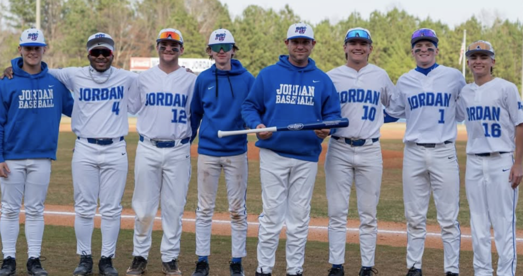 Mortimer Jordan's baseball coach holds a bat and smiles as he poses for a photo with some of his players on a baseball field.