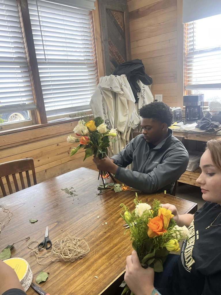 Students sit at a table inside a building and arrange flowers.