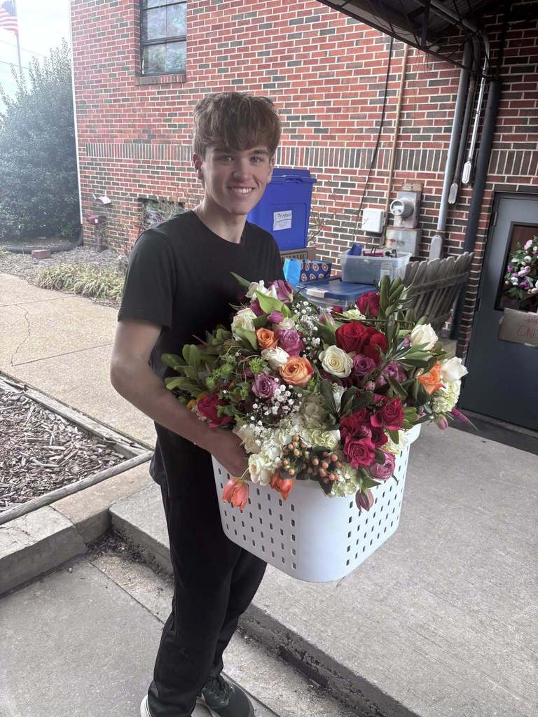 A student holds a basket of flowers and smiles for a photo while standing outside a building.