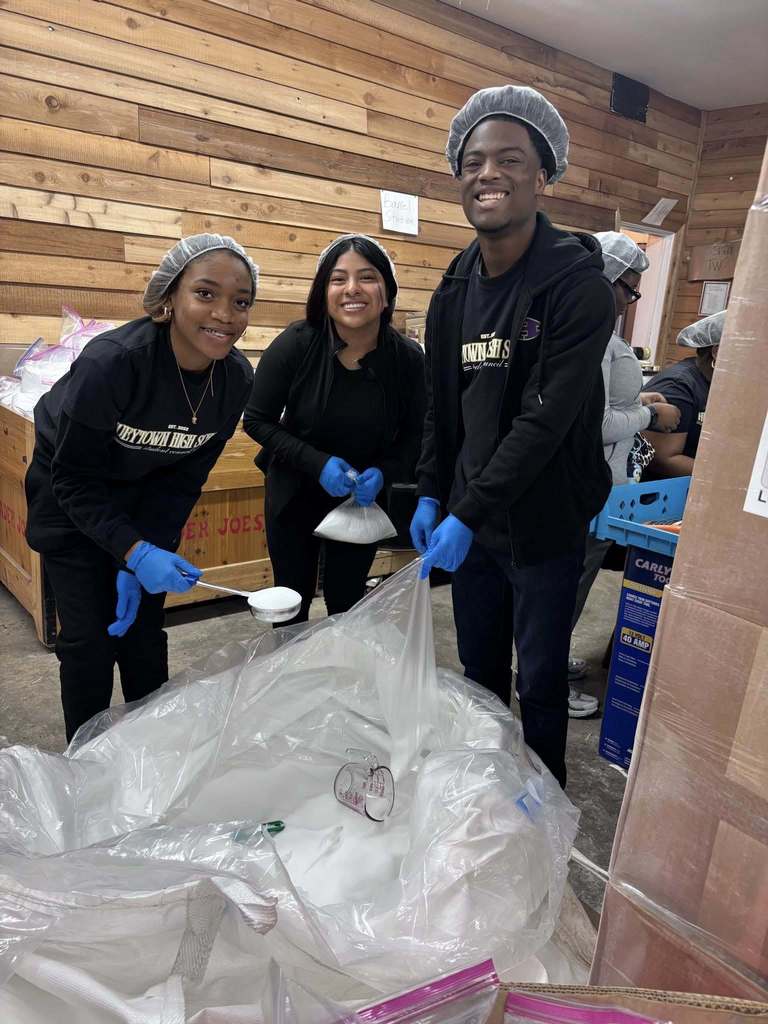 Students wearing all black outfits and hair nets scoop salt from a large bag and put it into smaller portions. The students smile for a photo while performing this task.