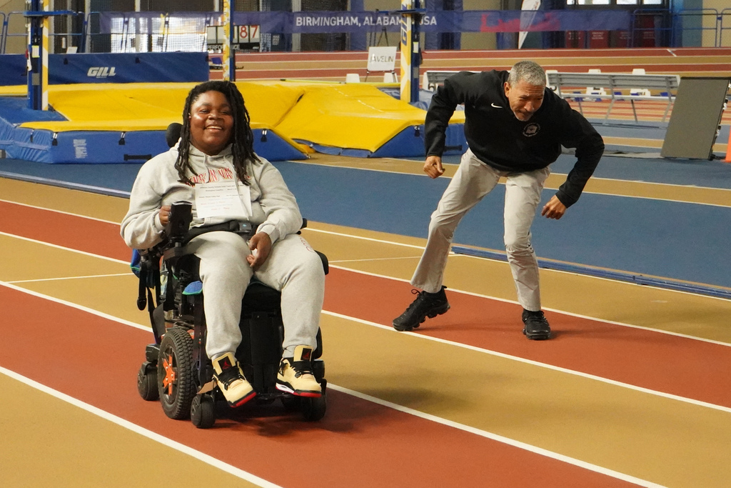 A student using a wheelchair races down an indoor track alongside an adult. 
