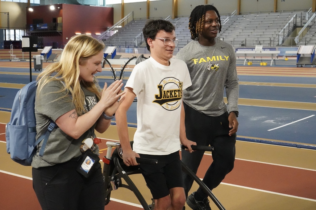 A McAdory student uses a walker to race down the track at the Birmingham CrossPlex. Another McAdory student walks next to him. On the other side of the student, a teacher walks and cheers him on.