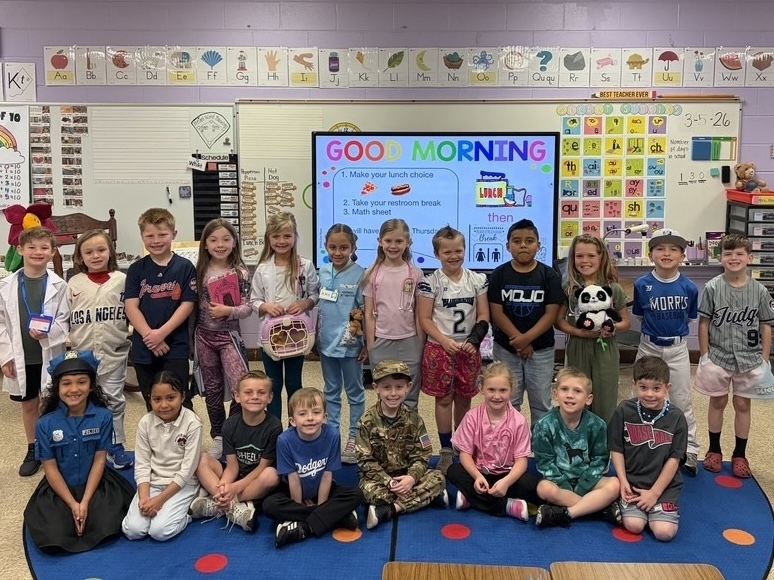 A class of students stand and sit in the room and smile for a photo. The students wear outfits inspired by what they want to be when they grow up. The costumes include health care workers, athletes, a police officers, an Army member, veterinarians , and athletes.