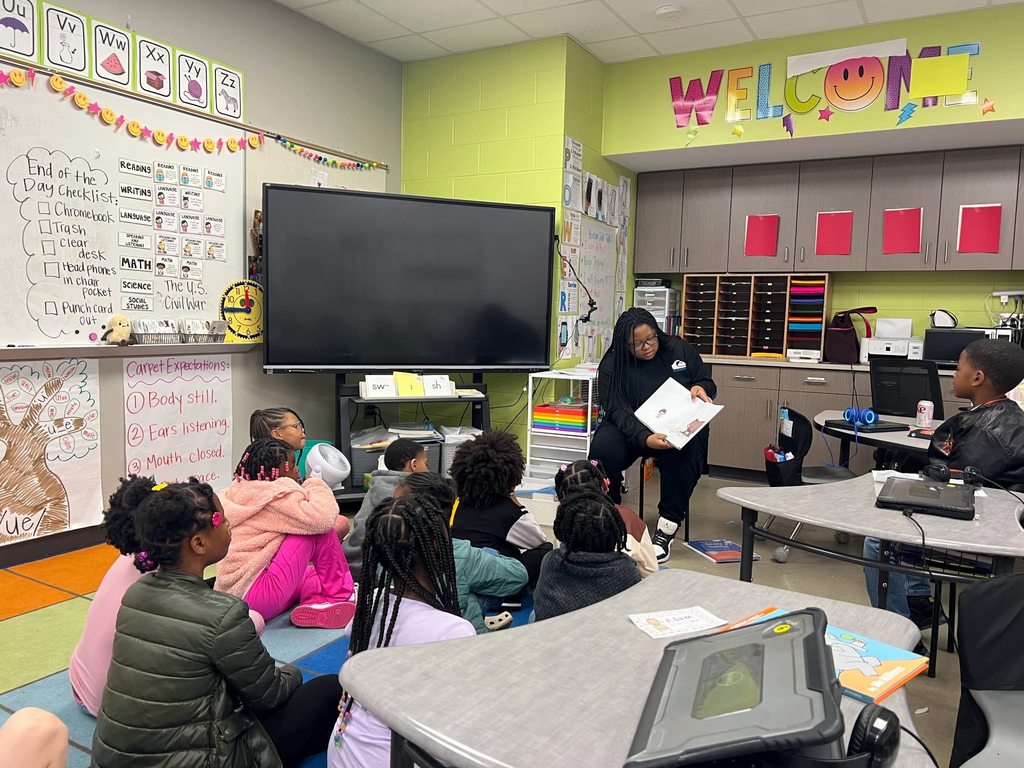 A person reads to a classroom full of students. The person sits in a chair while the students sit on a colorful rug or in a desk.