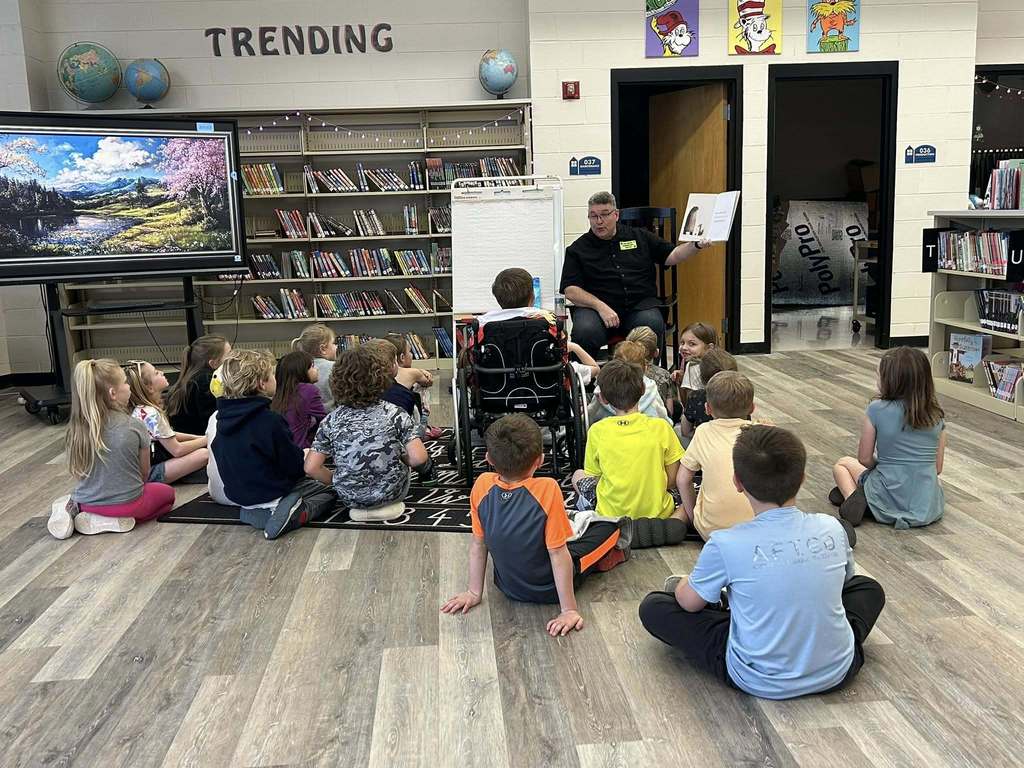 A man reads a book to students while they sit in the floor of a school library.