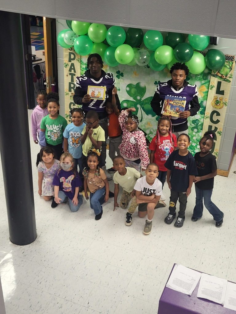 High School football players hold books and stand for a photo with young students inside a school.