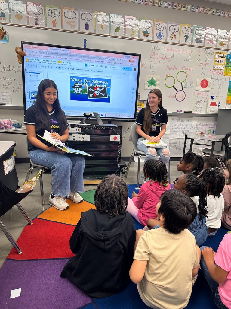 Two Hueytown High School students read to children in a classroom. The high schoolers sit in chairs and the children sit on a colorful rug.