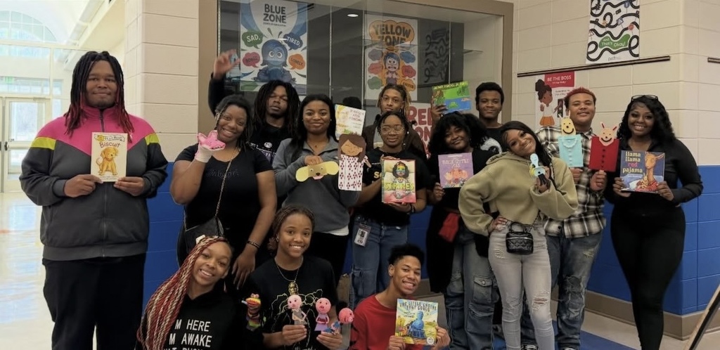 A group of high school students stand and sit together for a photo. They hold books and puppets.