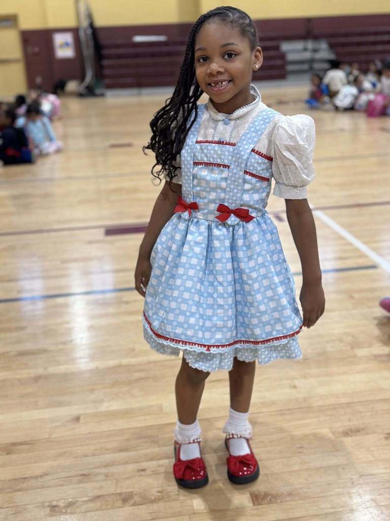 A student wearing a Dorothy costume smiles for a photo while standing in a school gym.